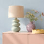 A blush console styled with a mint lamp, lilac vase, yellow scalloped tray, pastel art, books, and a small olive-green vase.
