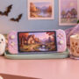 A pastel handheld game device, friendly ghost plush, mushroom lamp, pumpkin, warm drink, controller, and lavender blanket on a pink desk.