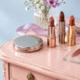Rose-brown and cocoa lipsticks, a silver compact, lip brush, lilac ribbon, and soft color swatches arranged on a blush-pink vanity.